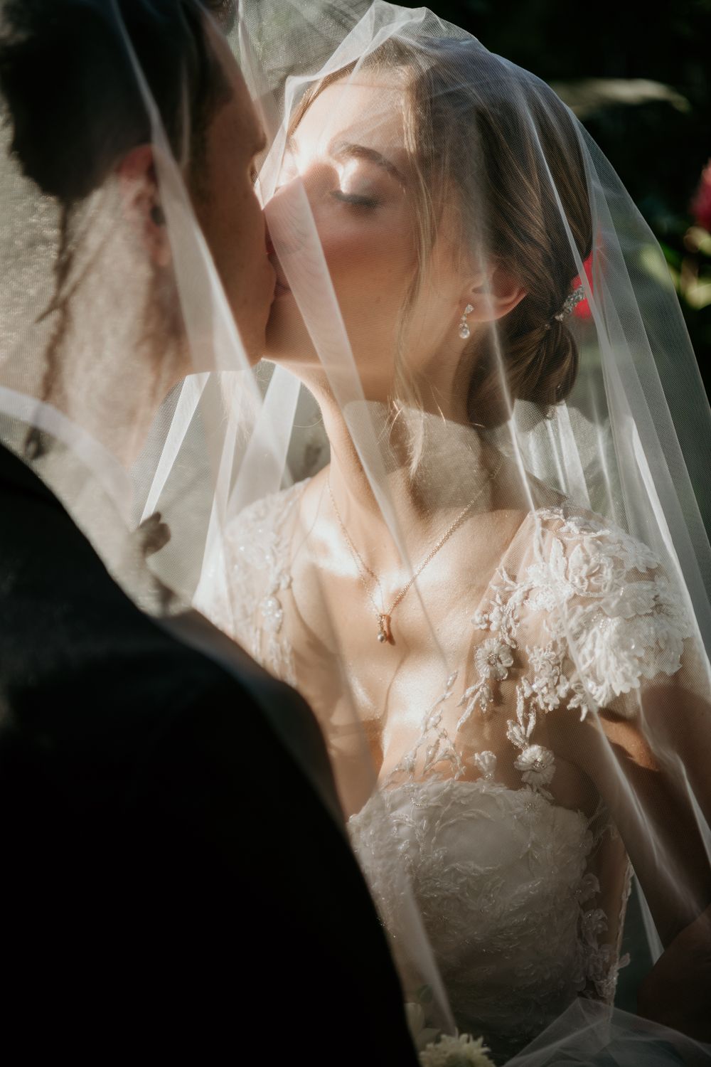 A romantic wedding moment captured through a delicate white veil with intricate lace details in soft natural lighting.