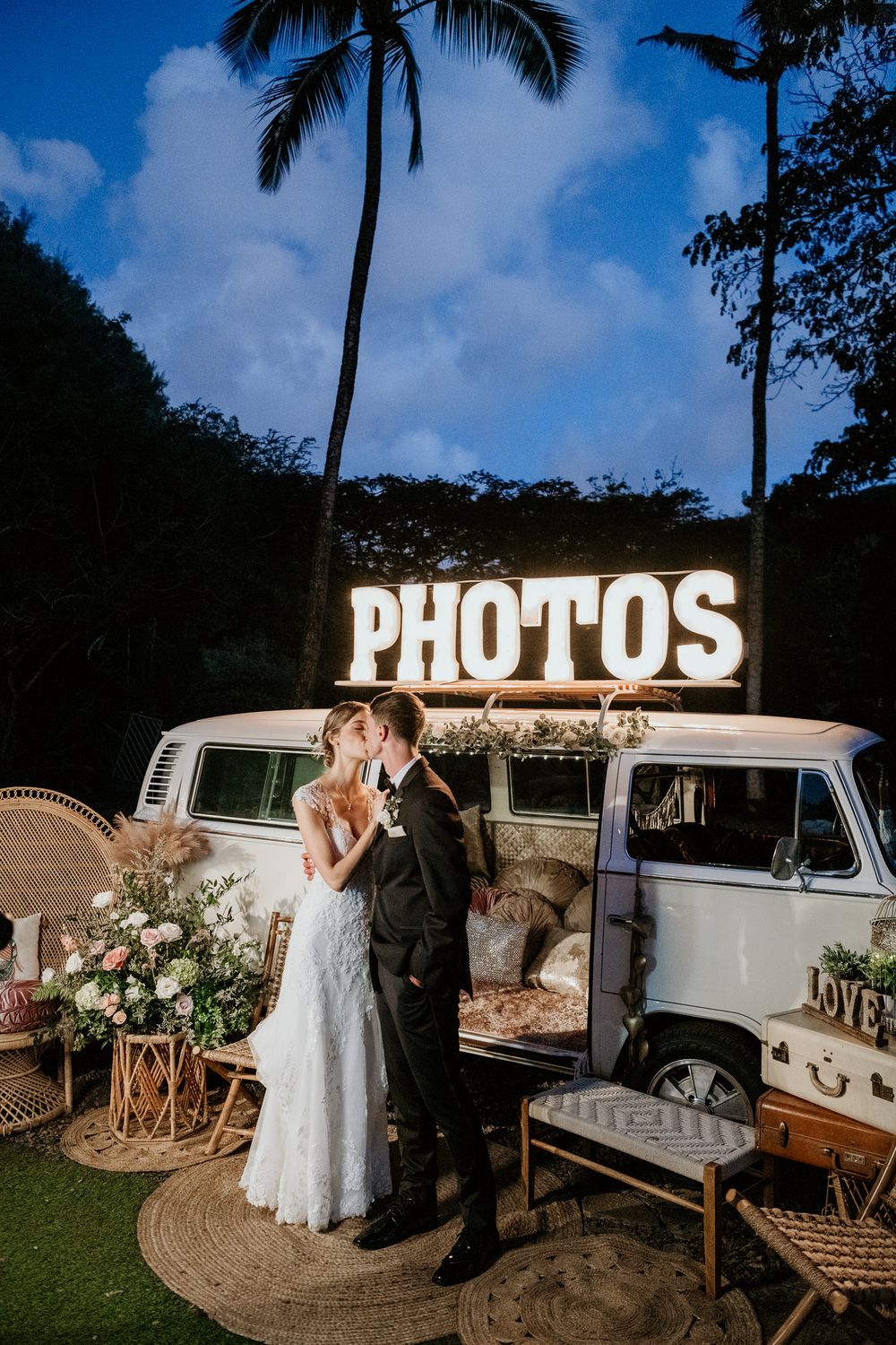 A vintage white VW bus serves as a photo booth backdrop with illuminated PHOTOS sign and tropical palm trees at dusk.