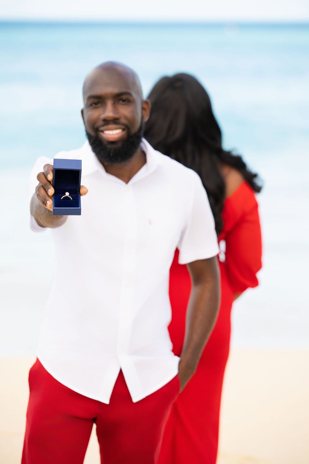 Person in white shirt and red pants holding blue ring box on beach.