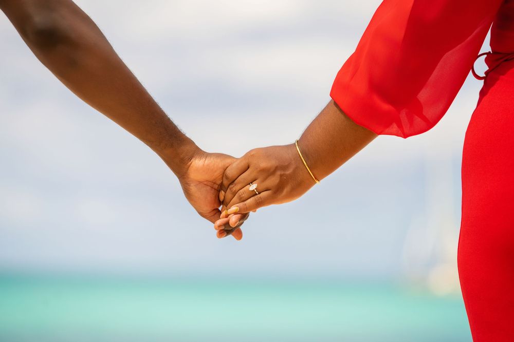 Close up of couple holding hands on beach with turquoise water in background.