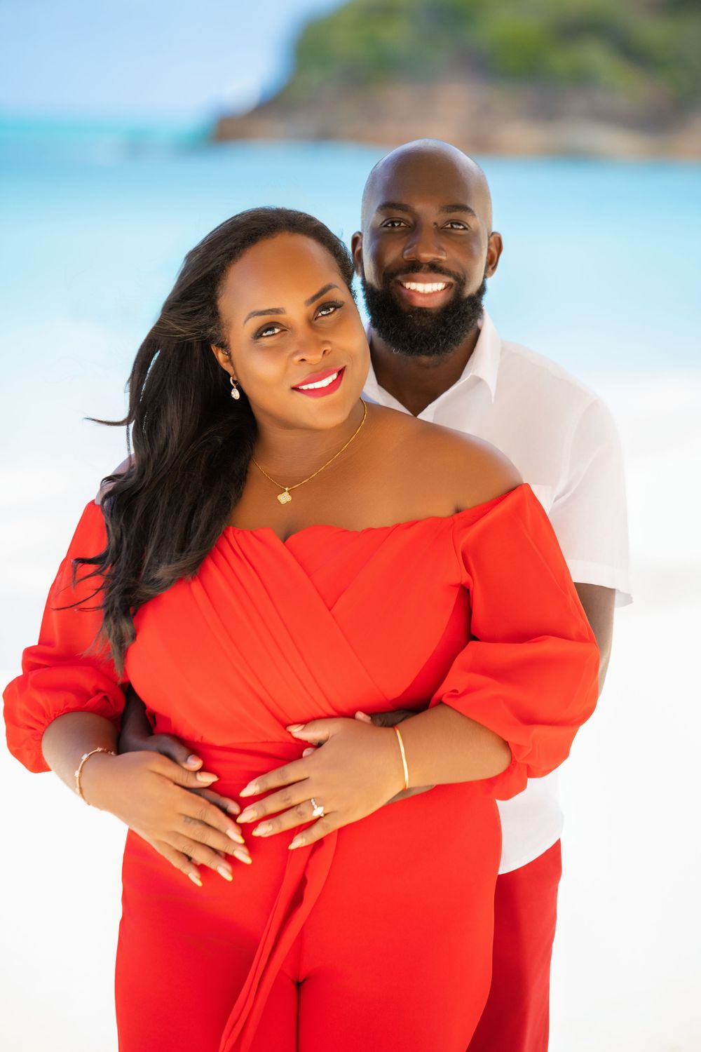 A romantic beach photo of a couple embracing, with her in a red dress and him in white against a tropical backdrop.