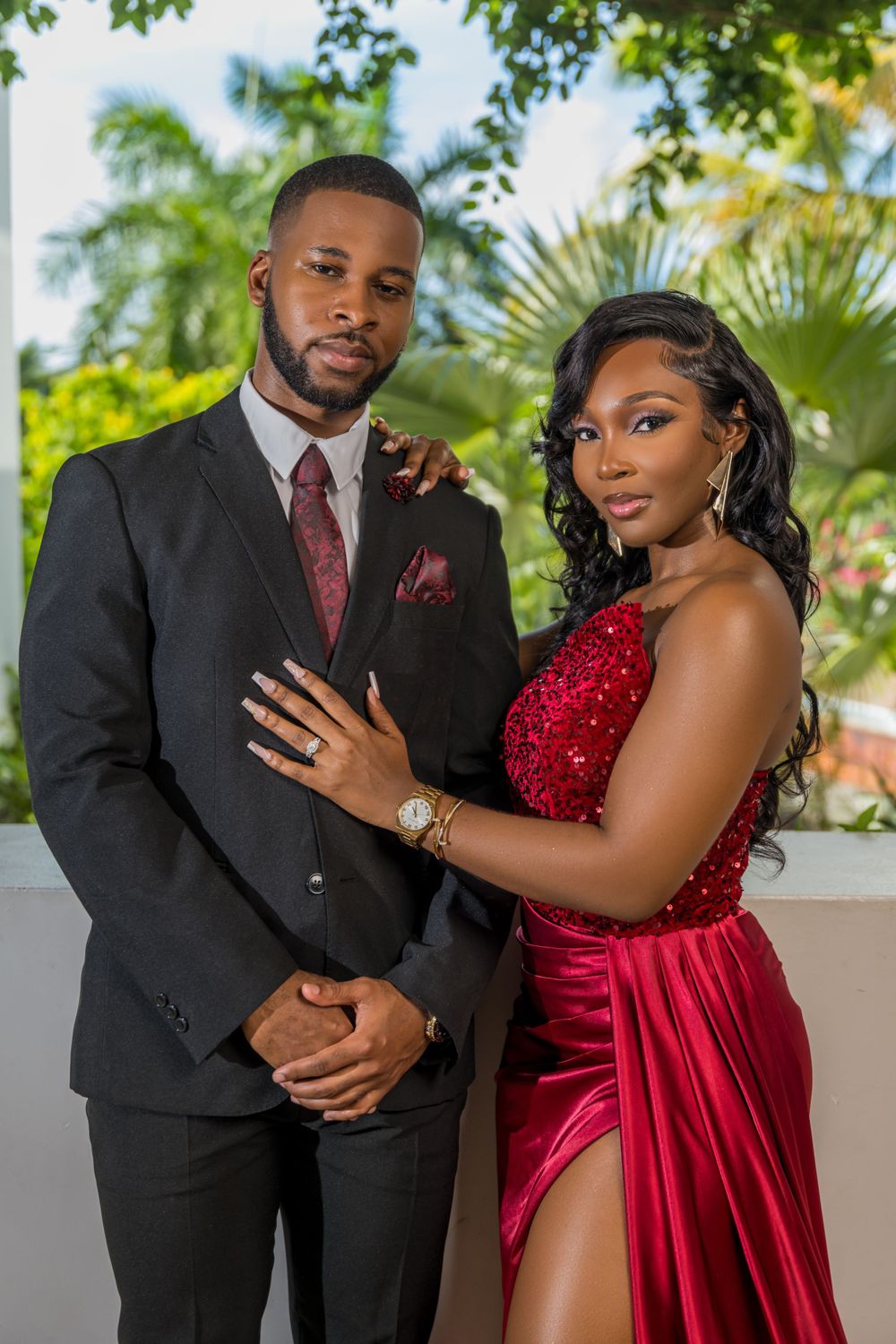 Elegant engagement photo of couple in black suit and red gown outdoors.