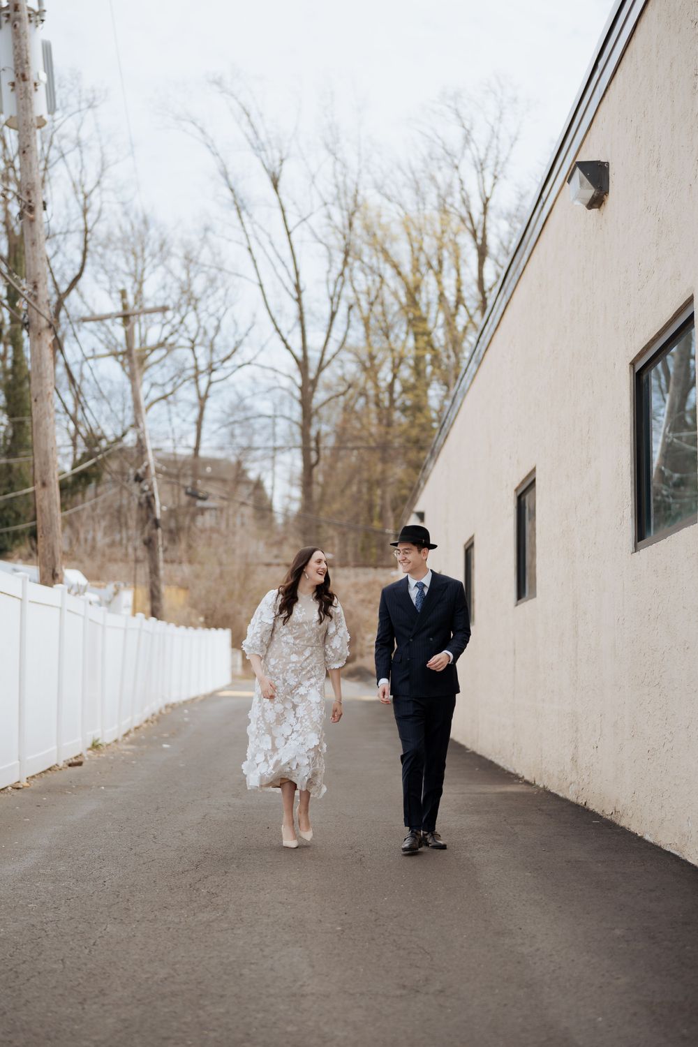 A couple walking together on a winter path beside a white fence and beige building.