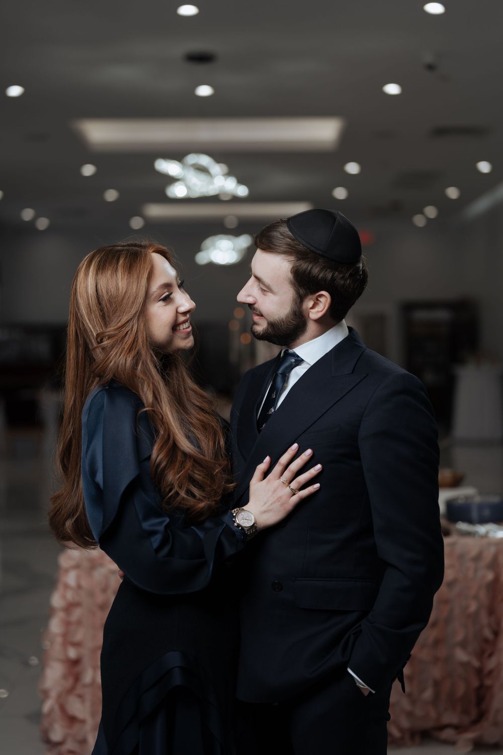 A romantic couple in formal attire share an intimate moment in an elegantly lit event space.