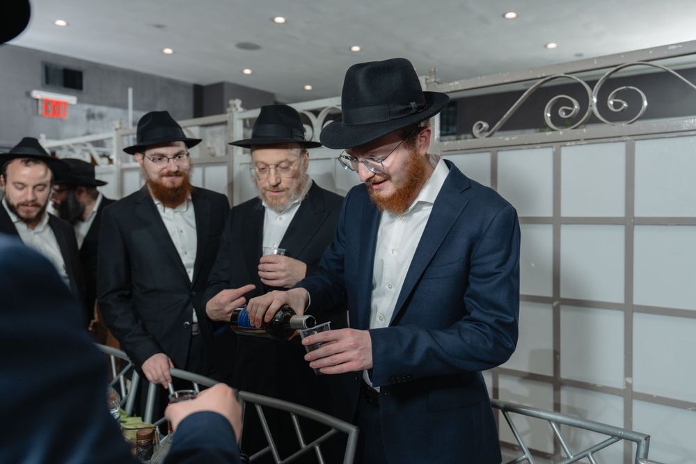 Group of Orthodox Jewish men in black suits and hats gathered in modern indoor setting.