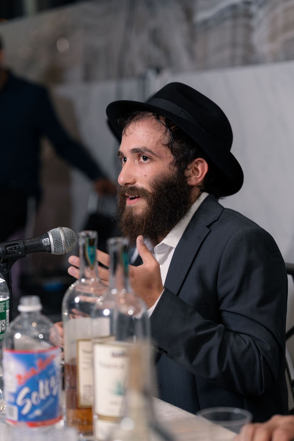 Orthodox Jewish speaker in black suit and hat smiles while speaking at microphone with drinks and bottles on table.