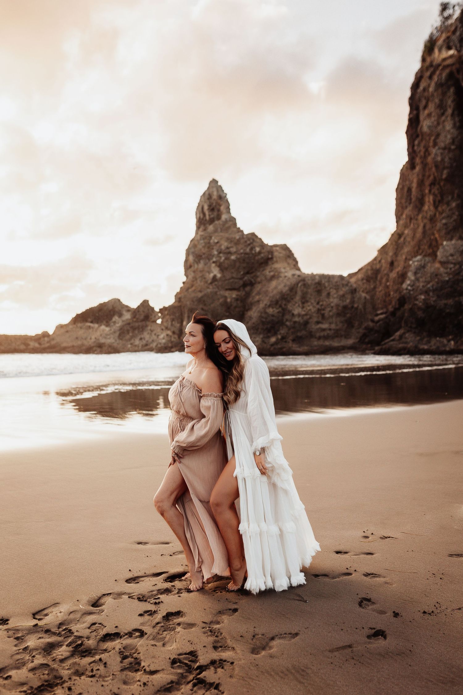 Two women in flowing dresses embrace on a scenic beach with dramatic rock formations at sunset.