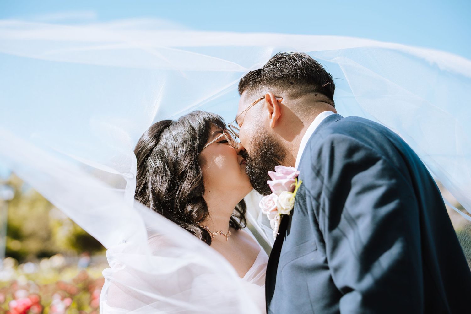 A romantic outdoor wedding moment as a couple embraces under a flowing white veil against a bright blue sky.
