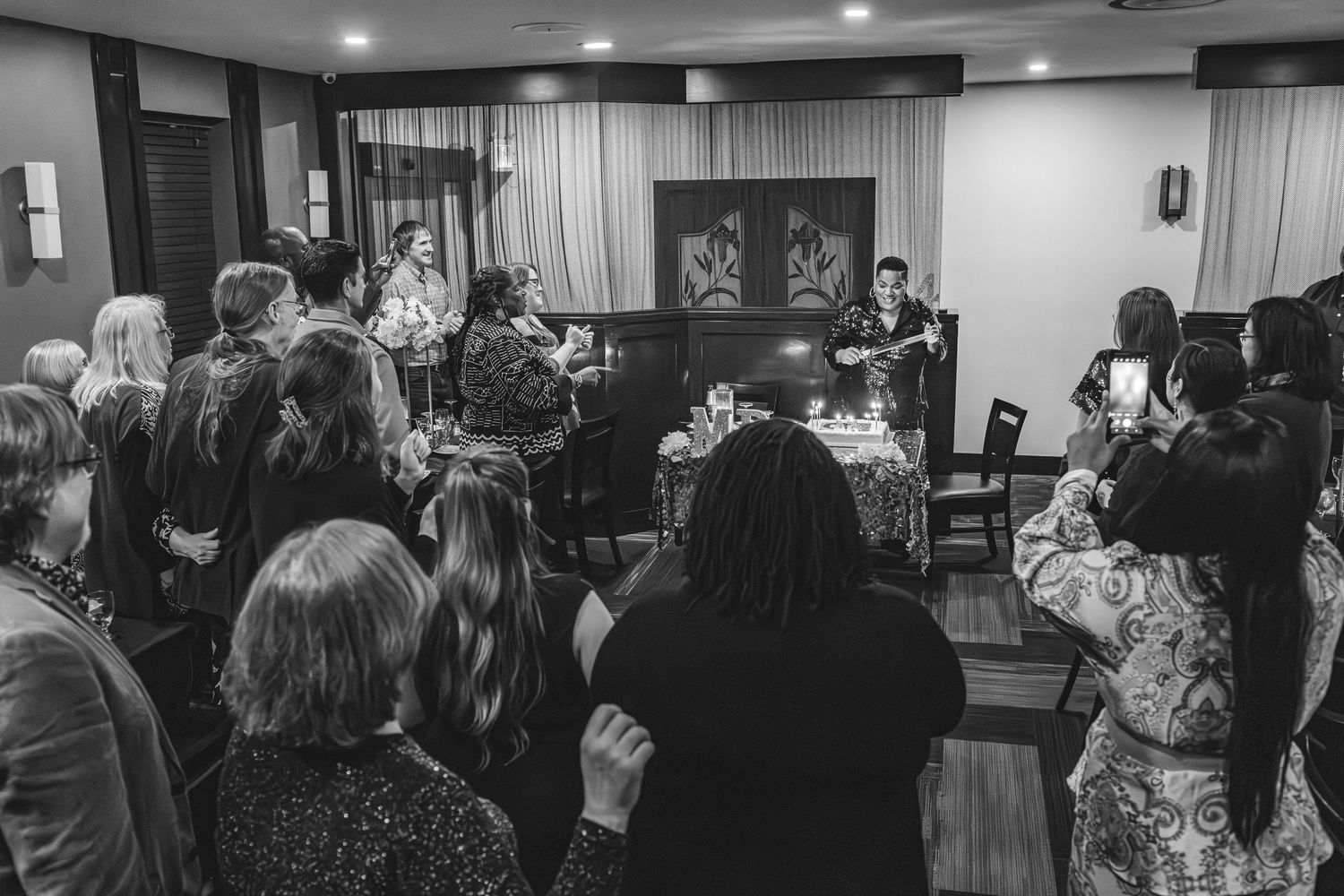 A black and white photo shows guests gathered around a piano performer at an intimate indoor venue.