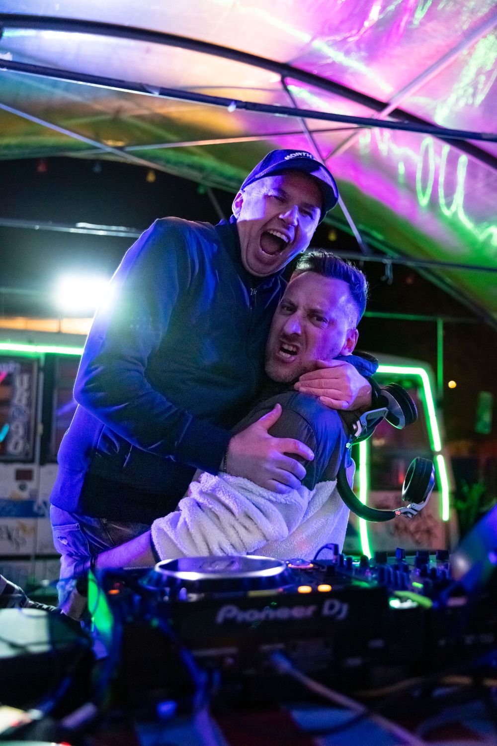 Two energetic DJs sharing a fun moment behind a Pioneer DJ setup under colorful neon lights at a nightclub party.