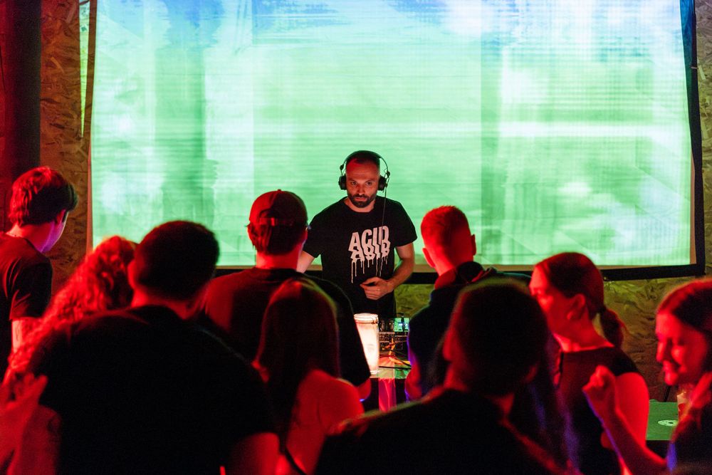 Performer stands on stage in front of a green projection screen while red lights illuminate the audience silhouettes.