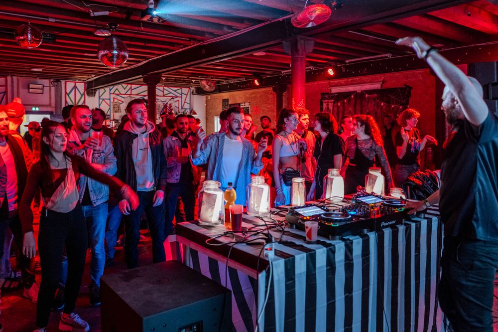 A crowd gathers around a display table under red lighting at an indoor nighttime event.