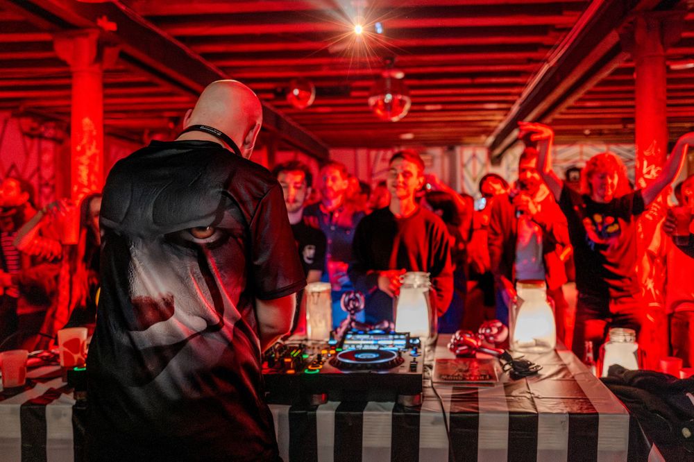 DJ performs at a nightclub bathed in red lighting while crowd dances behind the booth during an energetic party.