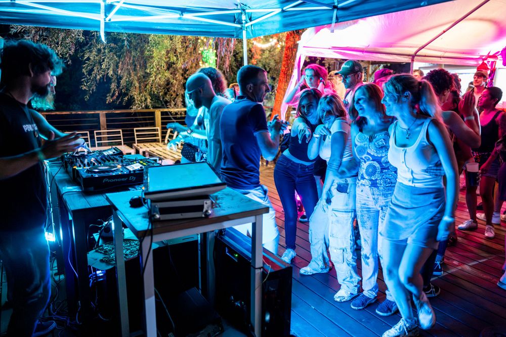 DJ performs under blue and red lighting at an outdoor event while attendees dance and enjoy the music.