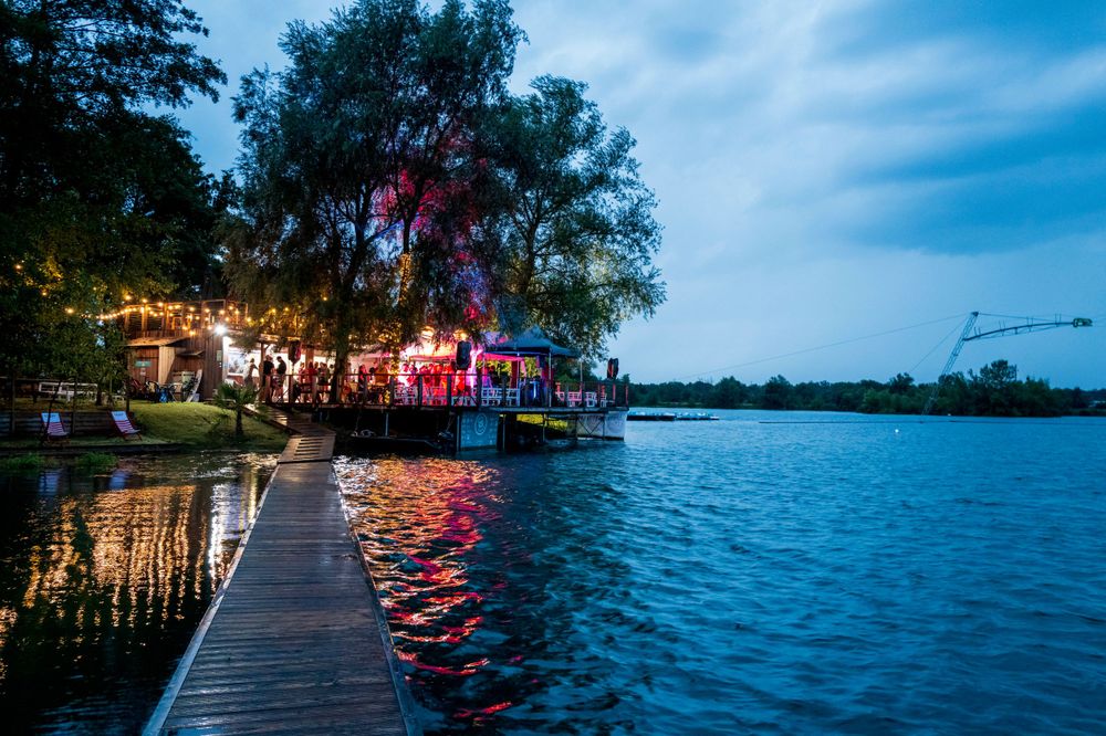Wooden dock leads to illuminated waterfront restaurant with string lights reflecting on calm lake at twilight.