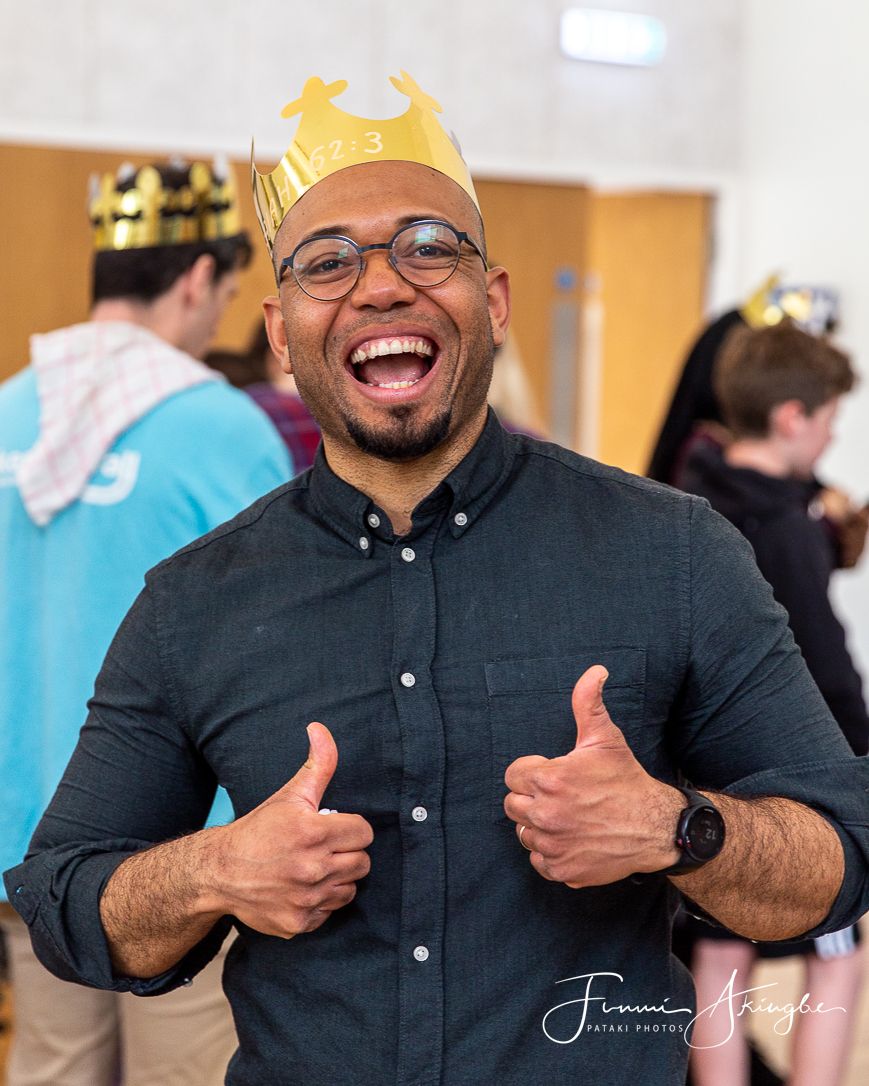 A joyful man wearing glasses and a paper crown gives enthusiastic thumbs up while wearing a dark button-down shirt.