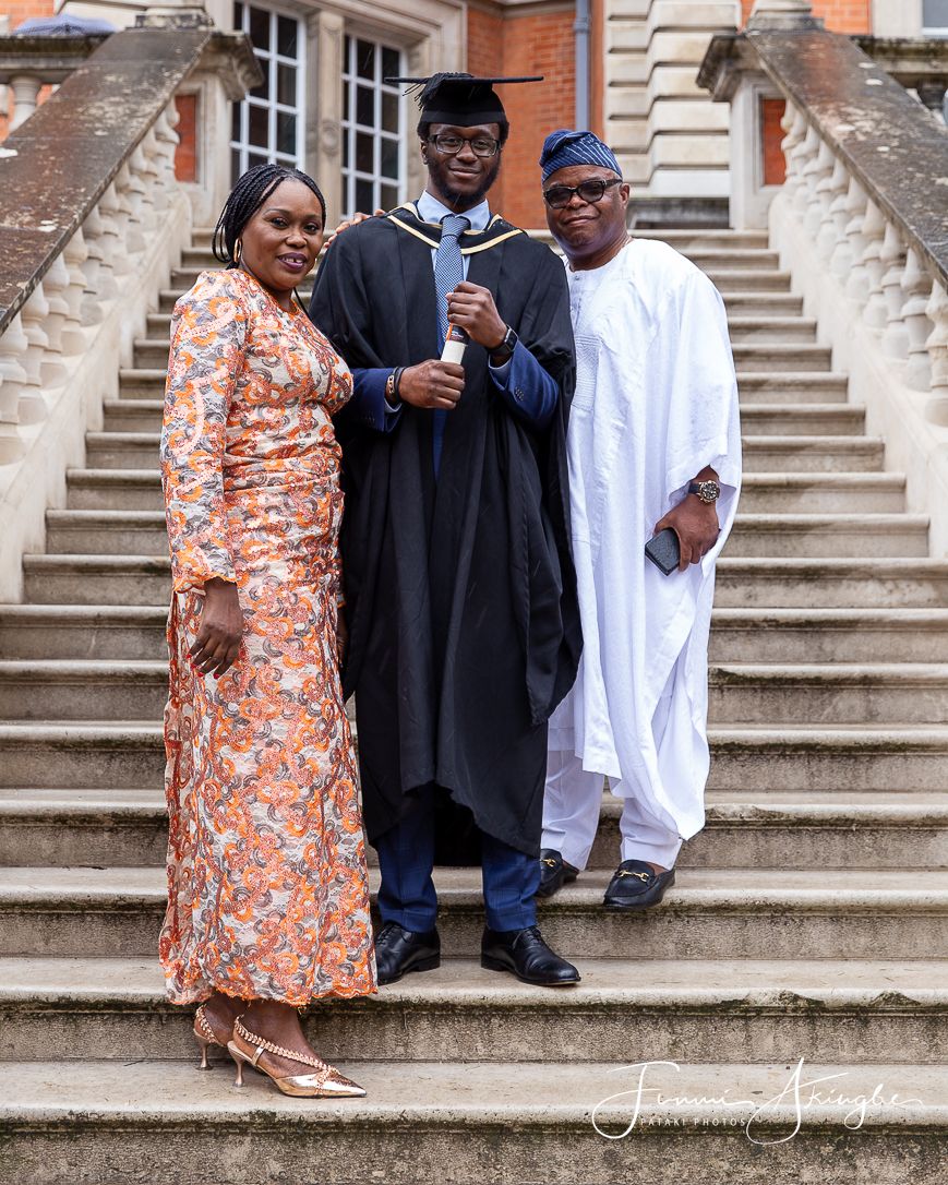 Family standing outside a university building with their son wearing his graduation gown and parents in traditional Nigerian attire.