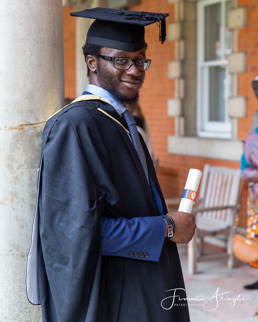 Graduate in academic regalia poses beside a column while holding a diploma on graduation day.