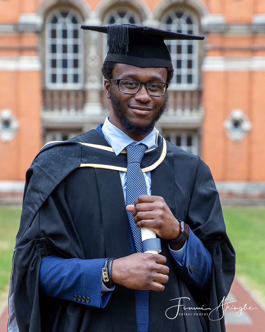 Graduate in black academic regalia and blue tie holds diploma in front of historic brick building with arched windows.