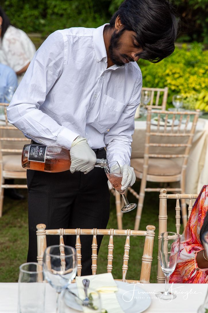 A server wearing a white dress shirt and black pants pours drinks at a garden party