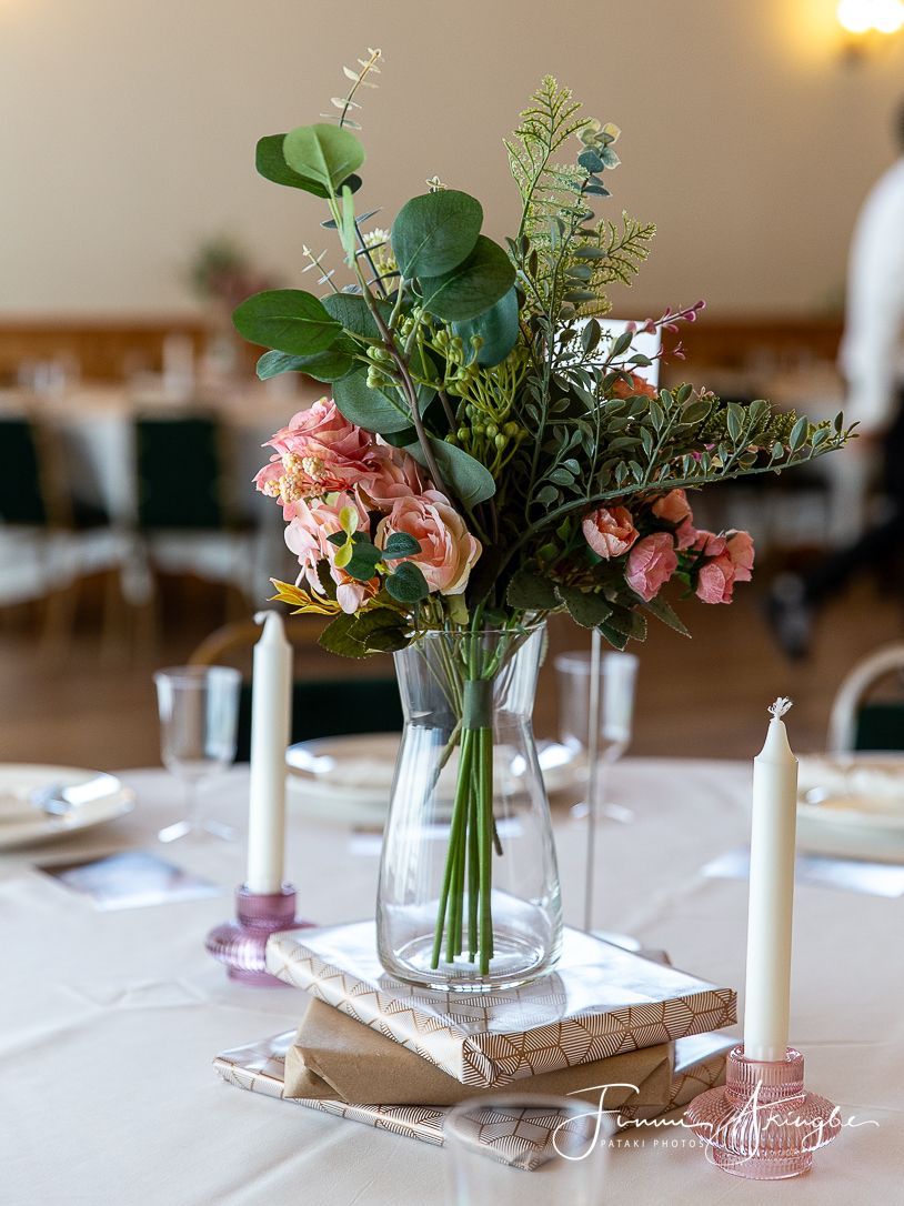 Gifts with floral centerpiece of pink roses and greenery in glass vase on white tablecloth with candles.