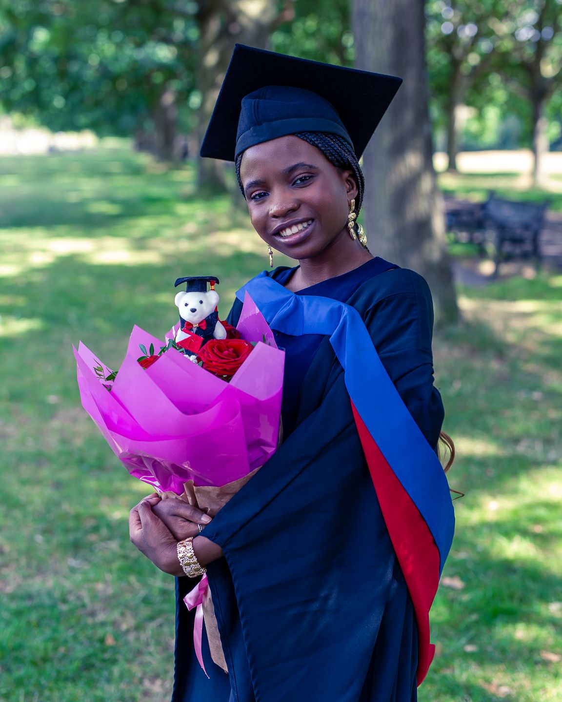A graduate in blue cap and gown holds a pink floral bouquet while standing in a sunny outdoor park setting.