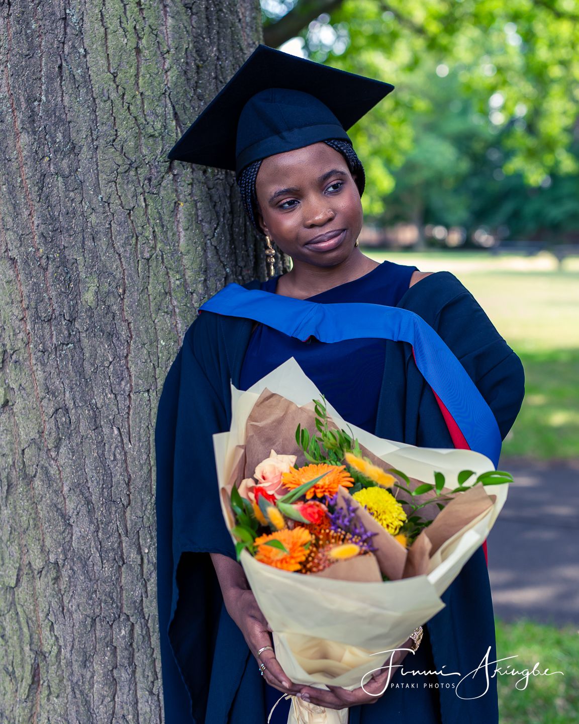 A graduate in academic cap and gown stands next to a tree holding a colourful bouquet of flowers in a park setting.