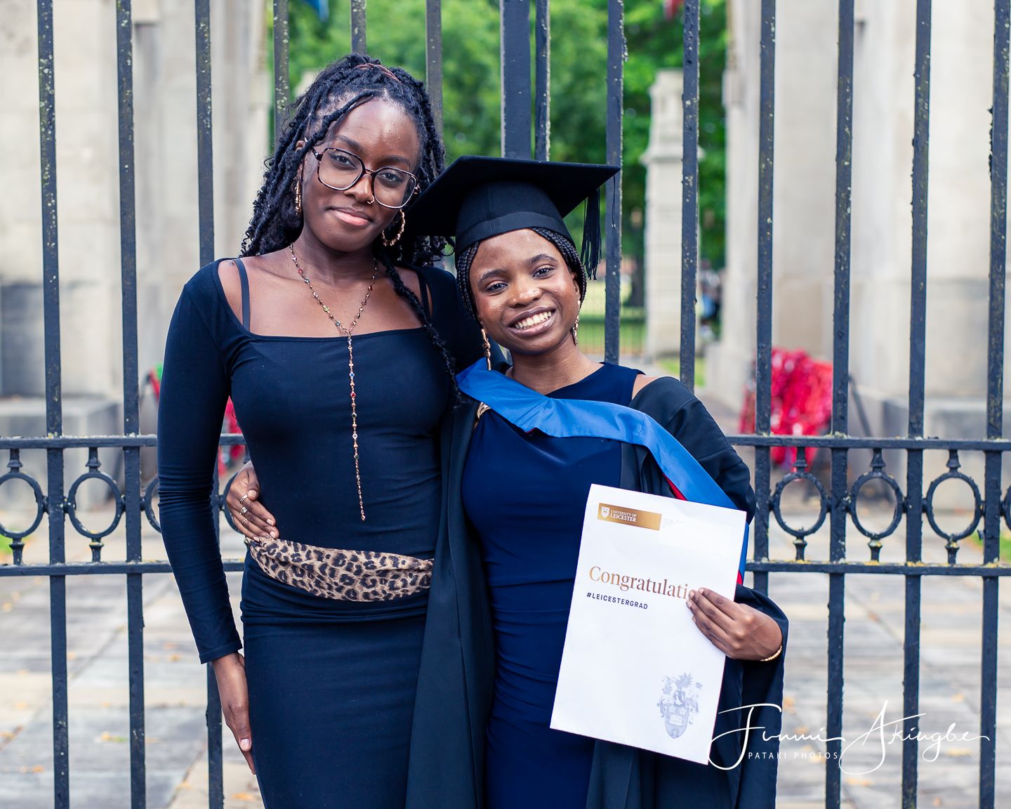 A young lady and female graduate wearing a cap and gown celebrating while holding a certificate and standing in front of wrought iron gates.