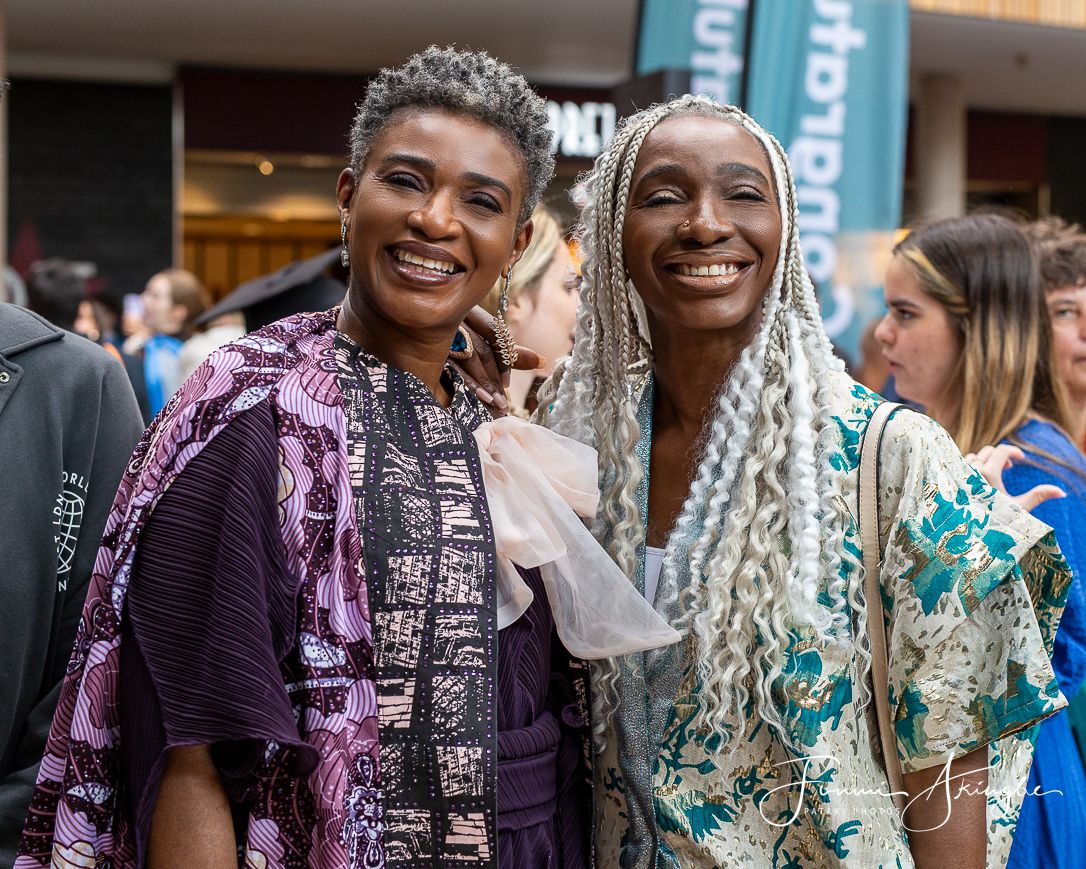 Two ladies wearing colourful Nigerian outfits smile warmly while standing together at an outdoor event.