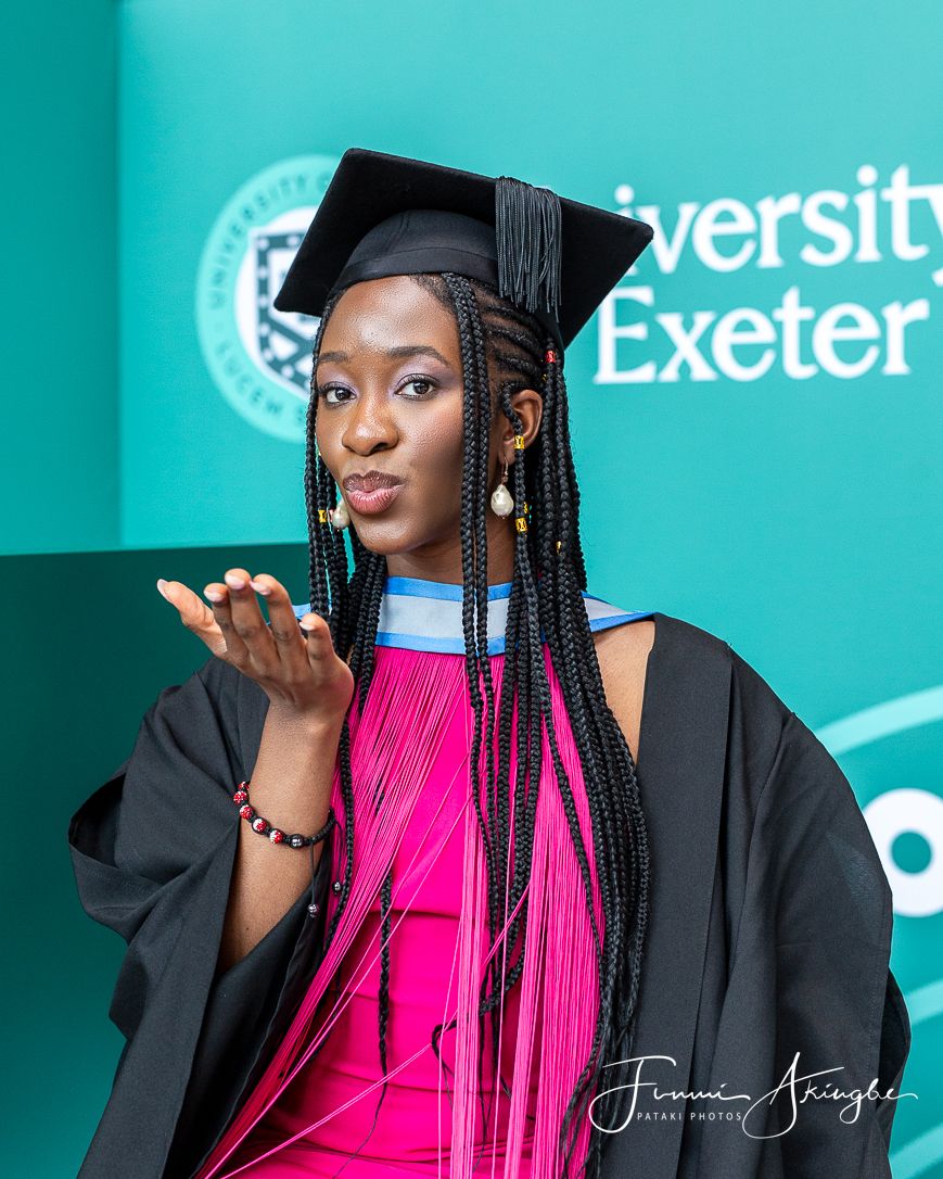 First-class graduate in academic regalia wearing pink dress and black graduation gown blows kiss at her graduation ceremony.