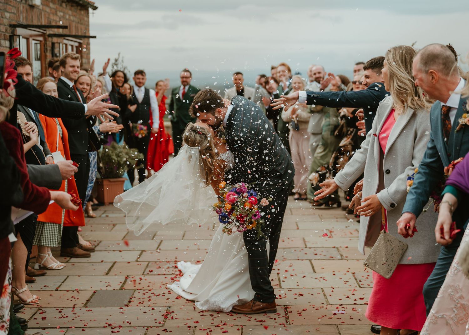 Wedding guests throw confetti over newlyweds on stone patio during joyous celebration.