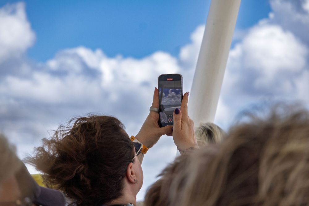 Crowd of people holding up phones to photograph something in the sky against a bright blue background.
