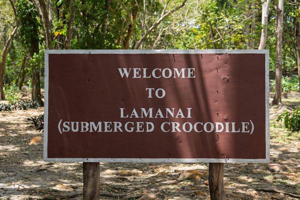 Brown wooden welcome sign for Lamanai archaeological site with tropical vegetation in background.