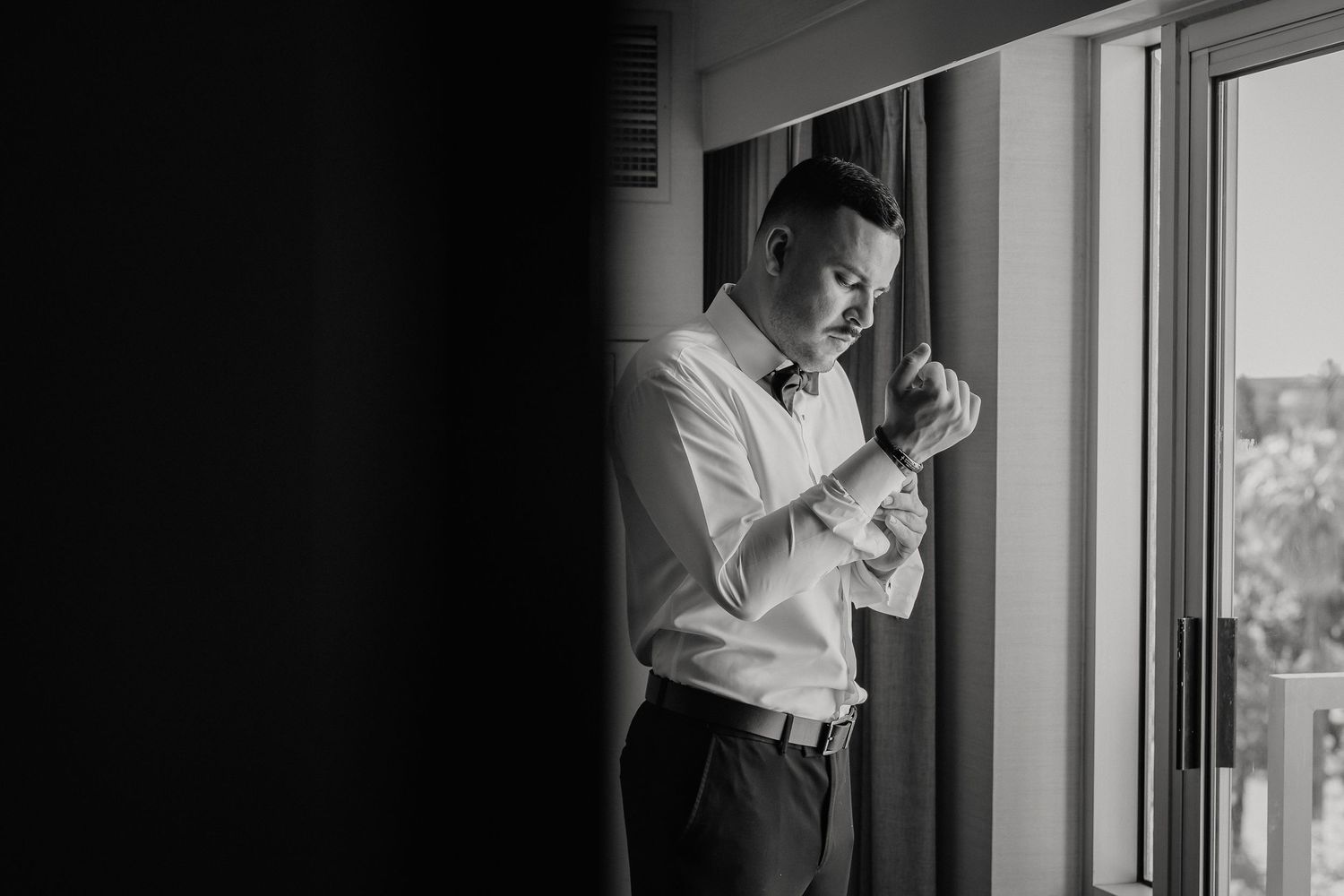 A person in formal attire adjusts their cuff while standing near a window in black and white photography.
