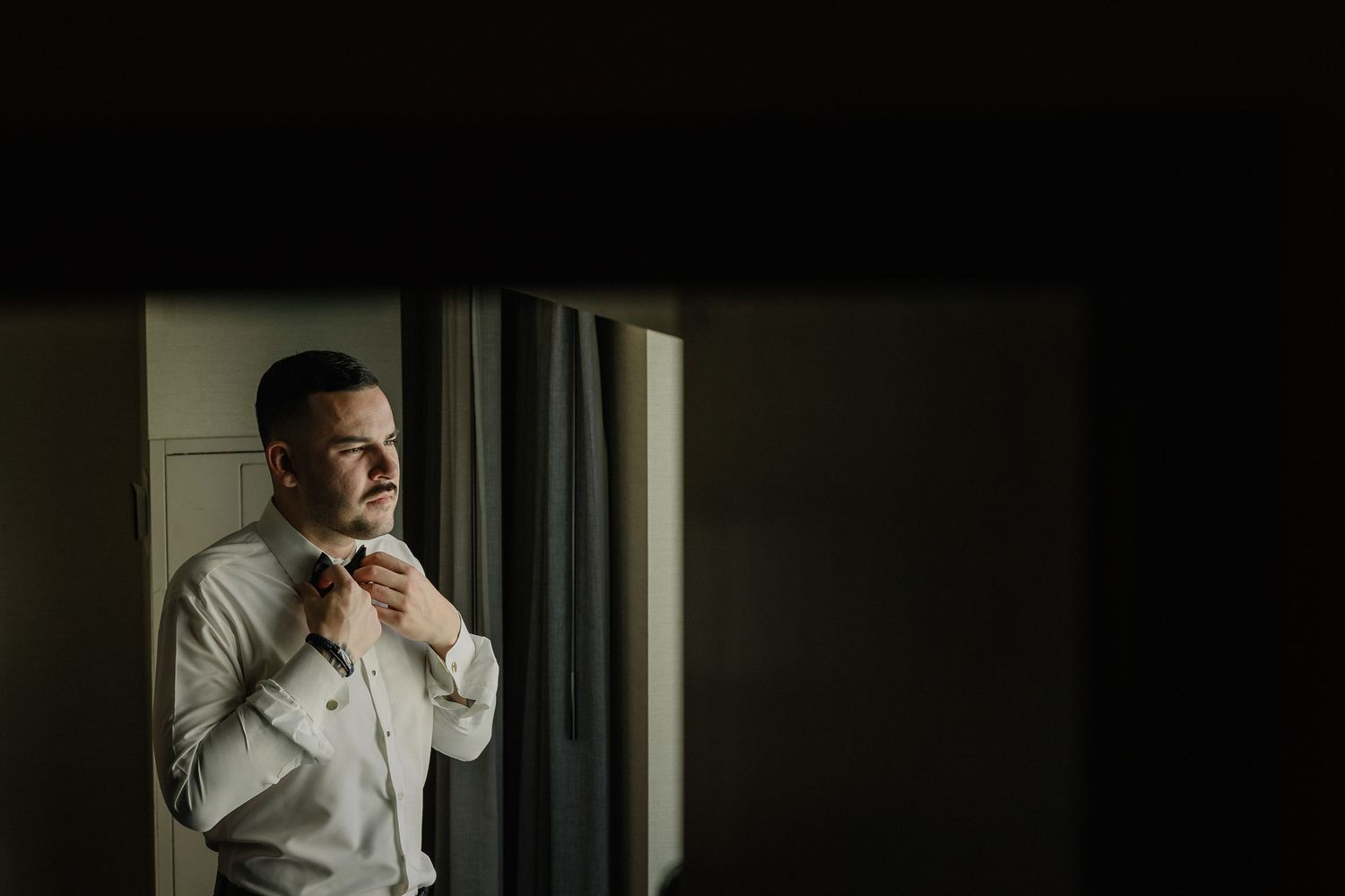 Groom adjusts his bowtie while standing by window in a moody, naturally lit scene.