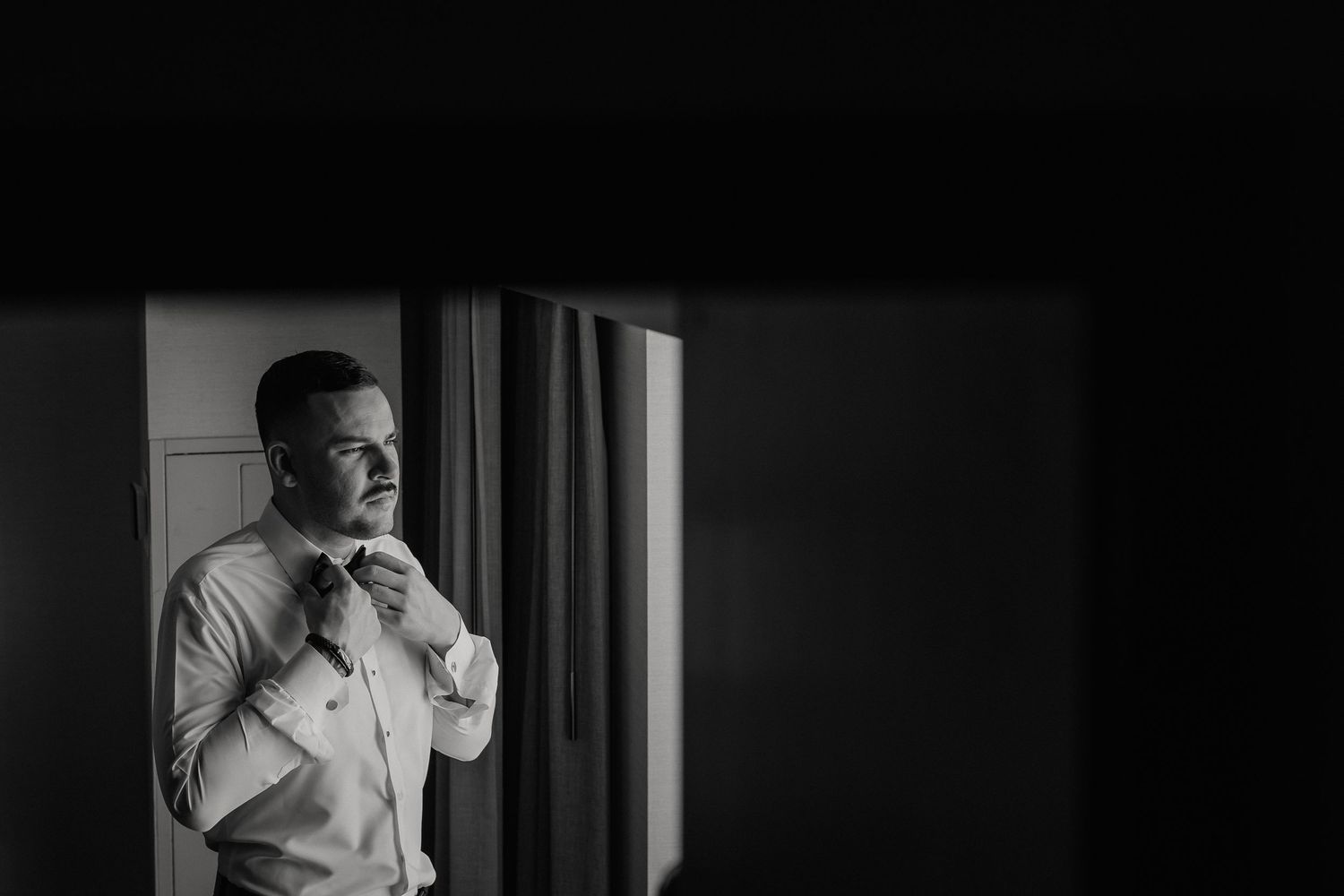 Black and white photo of a groom fixing his bowtie near a window in dramatic lighting.