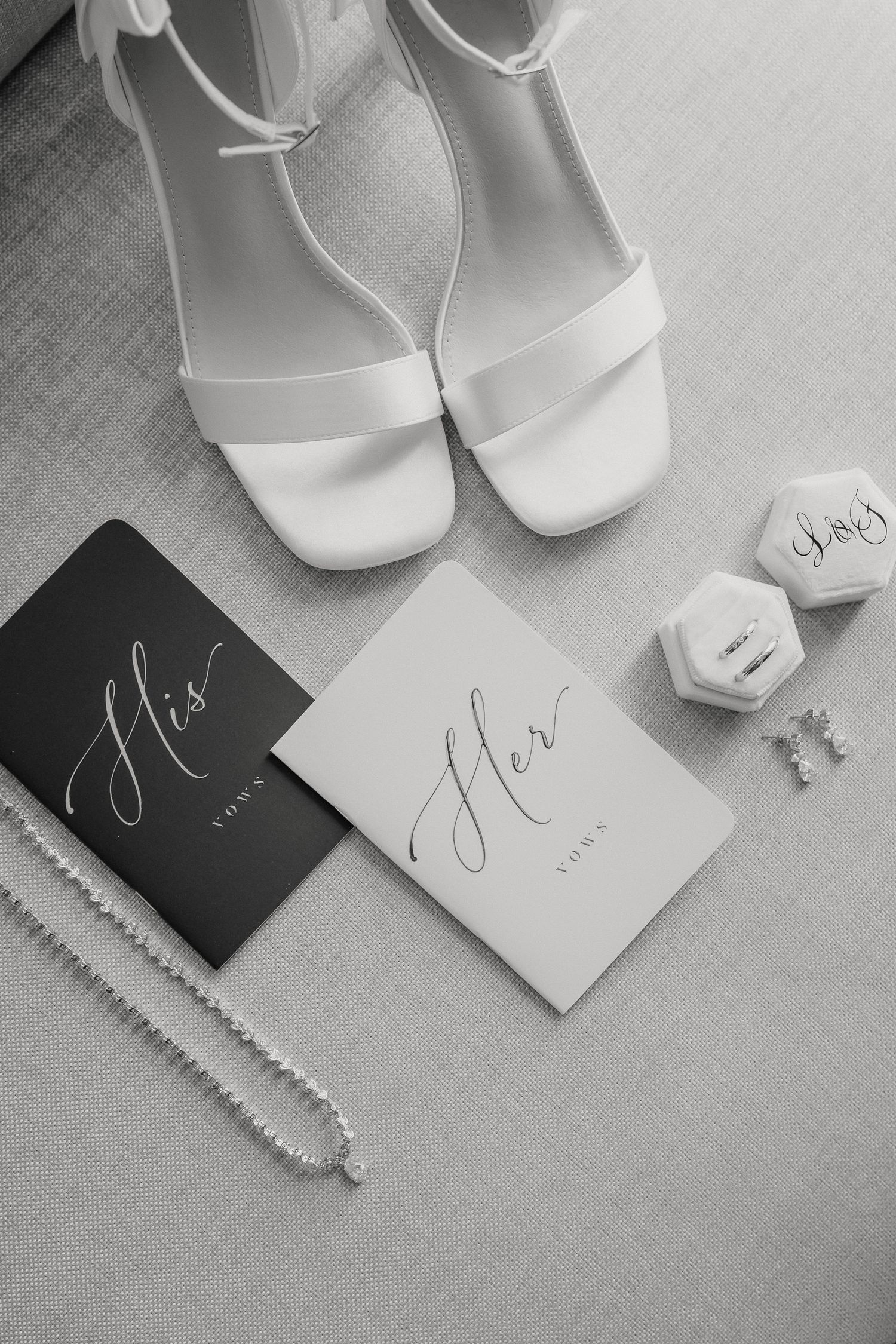 Black and white wedding detail photo showing white bridal shoes, 'her' cards, jewelry box and necklace on linen fabric.