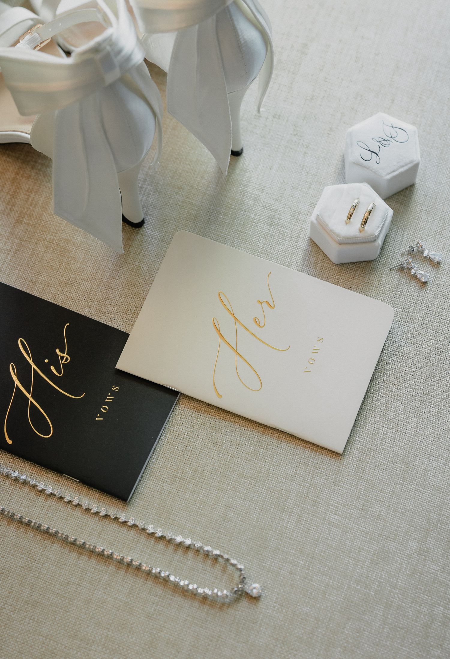 Wedding accessories including white heels, pearl necklace, ring box and black and gold vows books arranged on linen fabric.