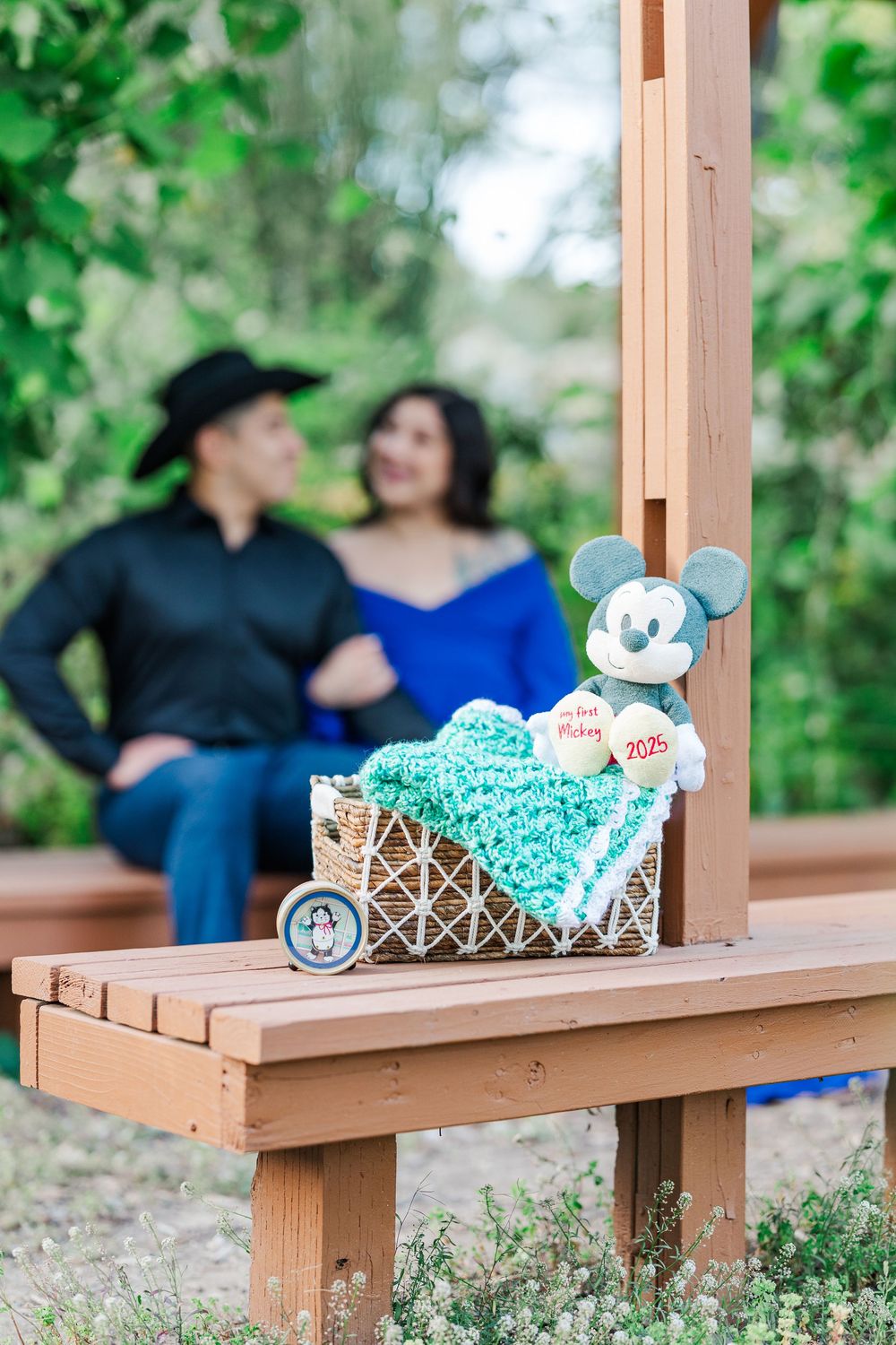 Baby nursery themed decor with Mickey Mouse and teal blankets displayed on wooden bench with couple posing in background.