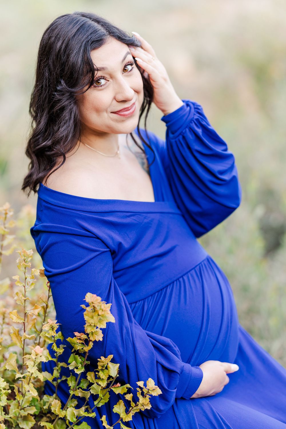 An expectant mother in a flowing royal blue dress poses outdoors next to yellow wildflowers during a maternity photoshoot.