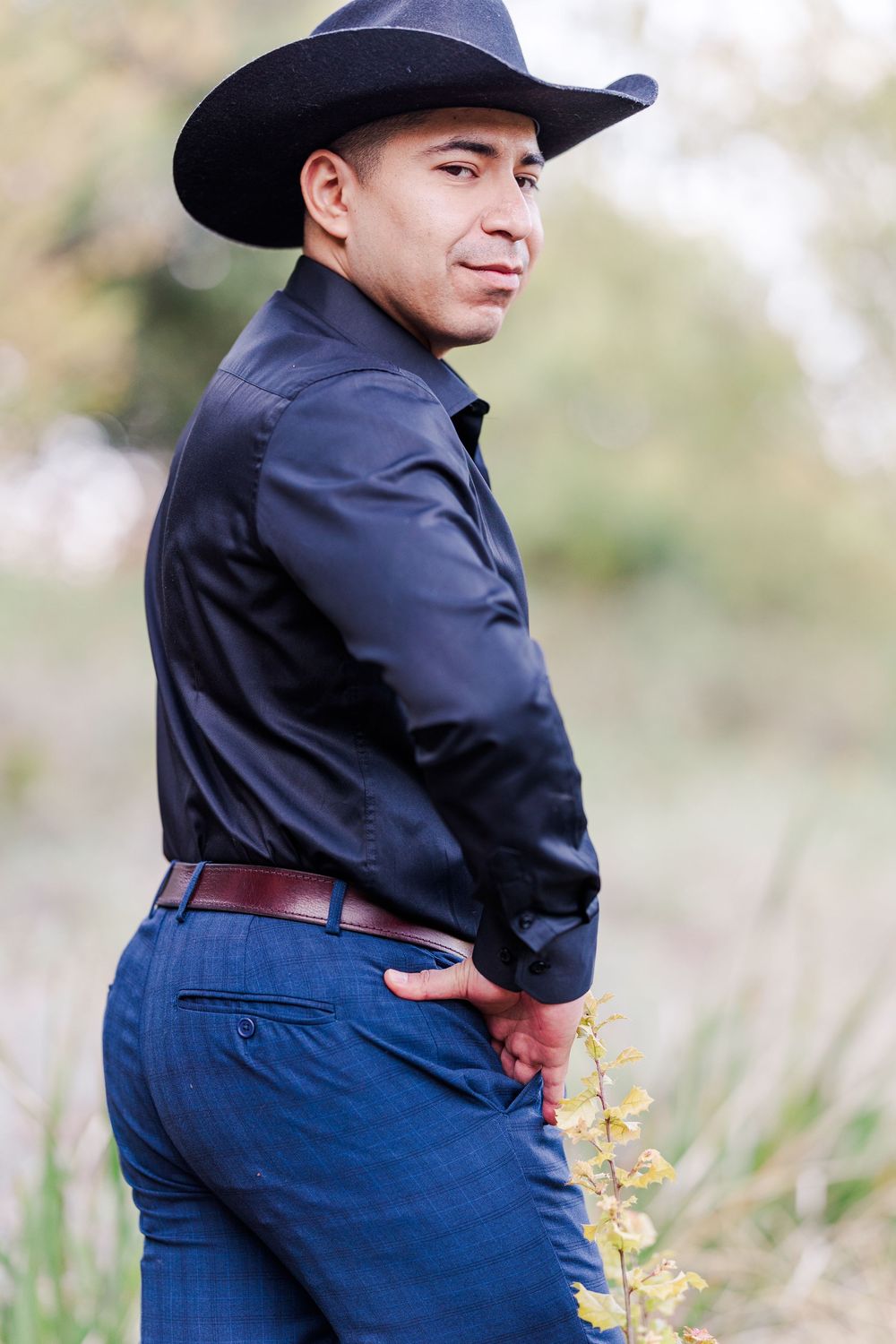 A person in a black cowboy hat and dark jacket poses outdoors in western attire with blue pants.