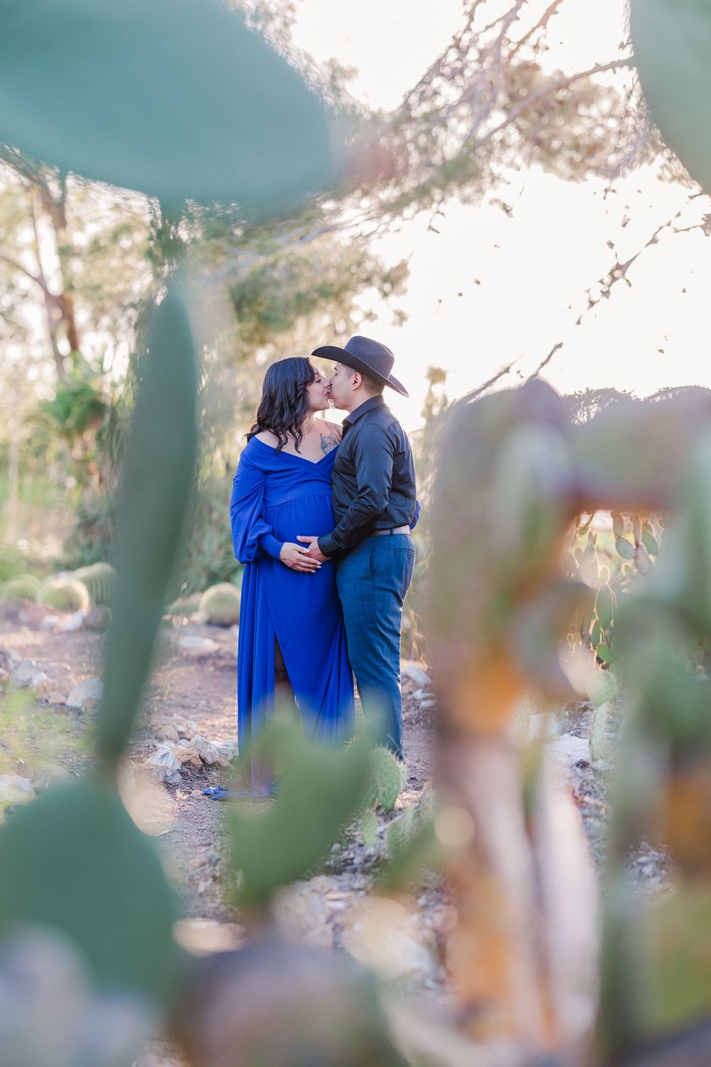 Two people in blue and black attire share an intimate moment on a outdoor desert trail with blurred foreground foliage.