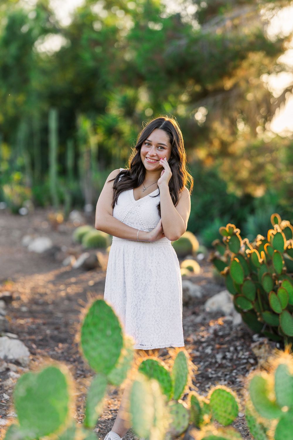 Woman in white dress poses outdoors among desert cacti and greenery at sunset.