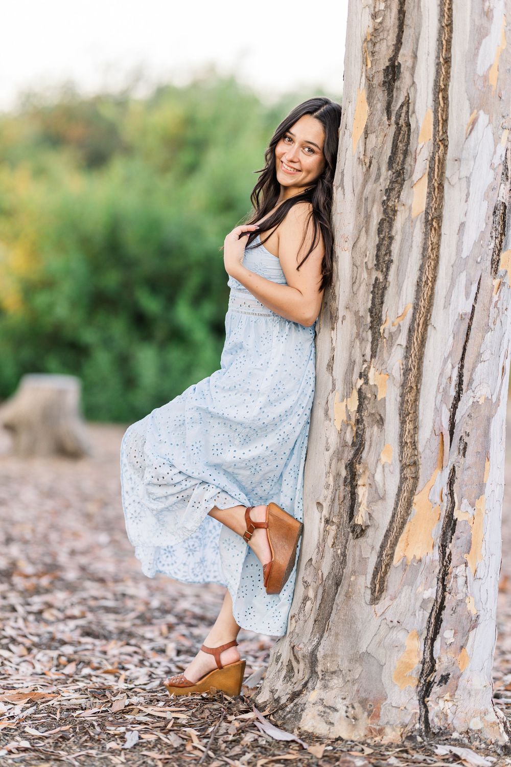 A woman in a flowing light blue dress poses against a rustic textured wall in a natural outdoor setting.
