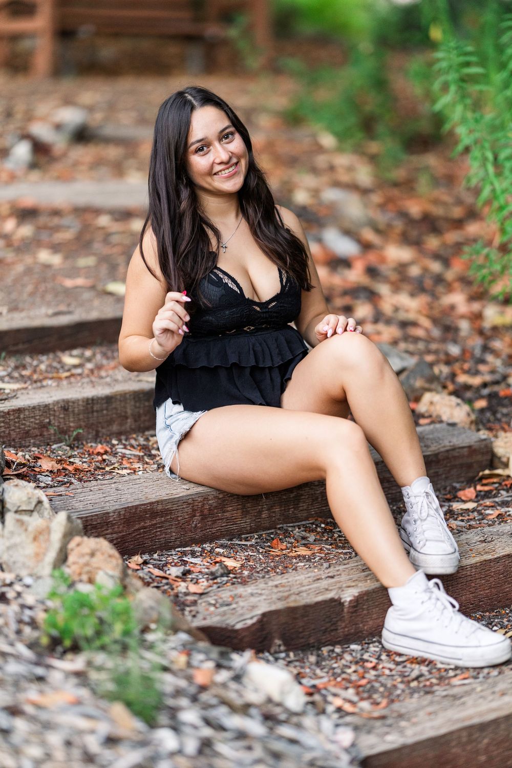 Outdoor portrait on rustic wooden steps surrounded by fallen leaves, wearing black top and white sneakers in natural setting.