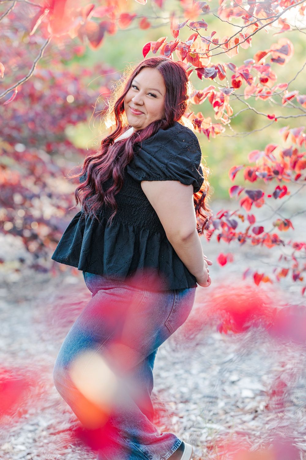A woman in a black dress poses beneath vibrant red autumn leaves creating a dreamy outdoor portrait.