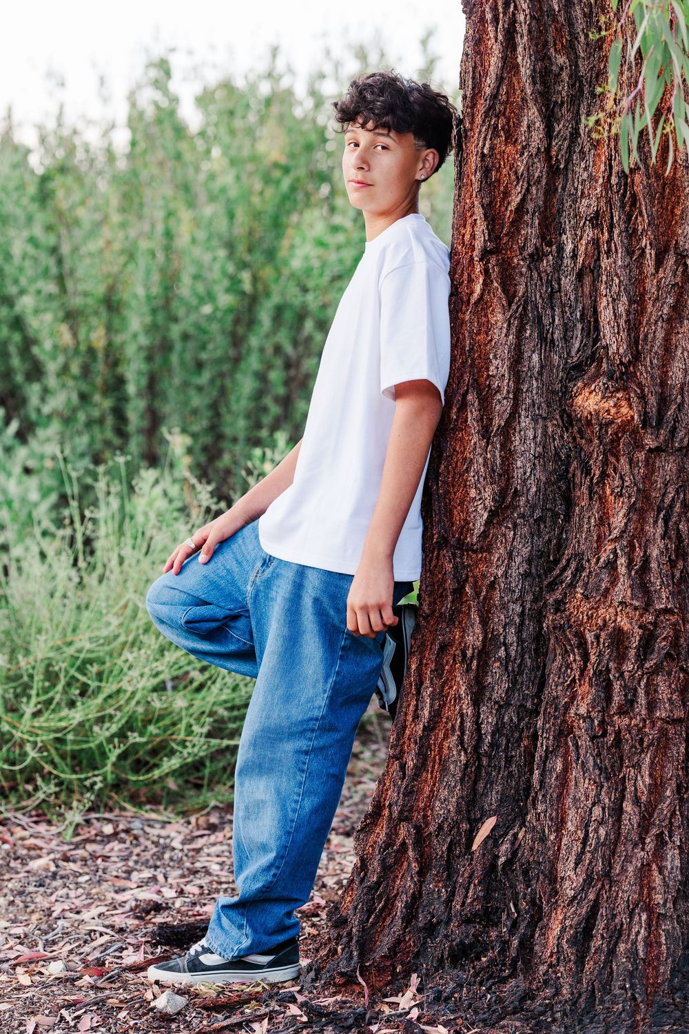 Child in jeans and white shirt leaning against large tree trunk with grassy foliage in background.