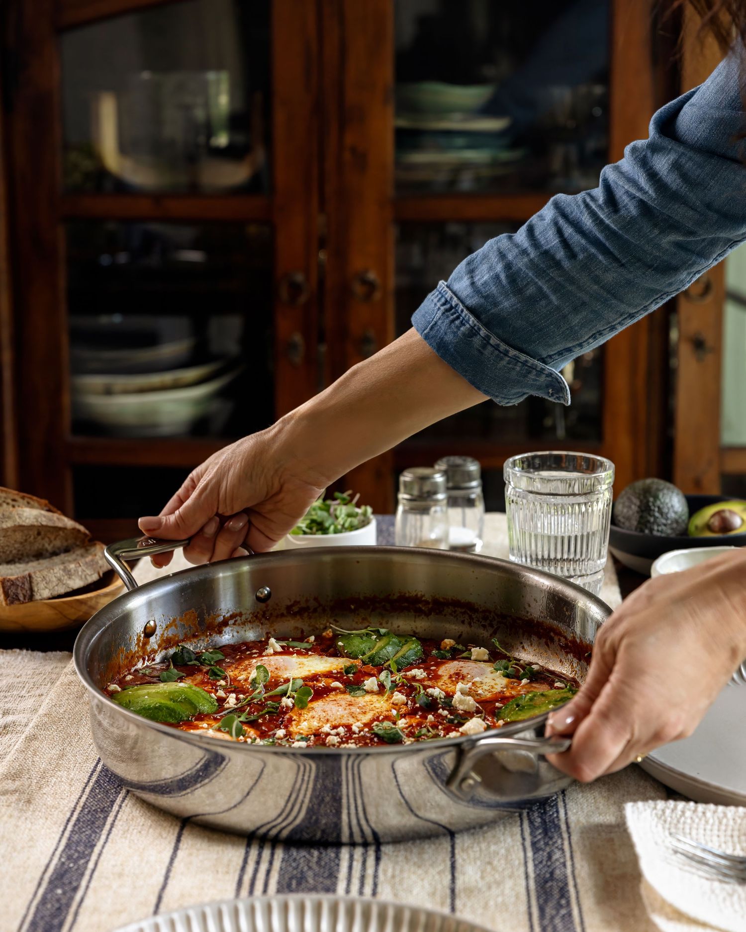 Hands reach across a rustic dining table to serve a skillet of shakshuka All Clad Mother of All pans with eggs and fresh herbs.