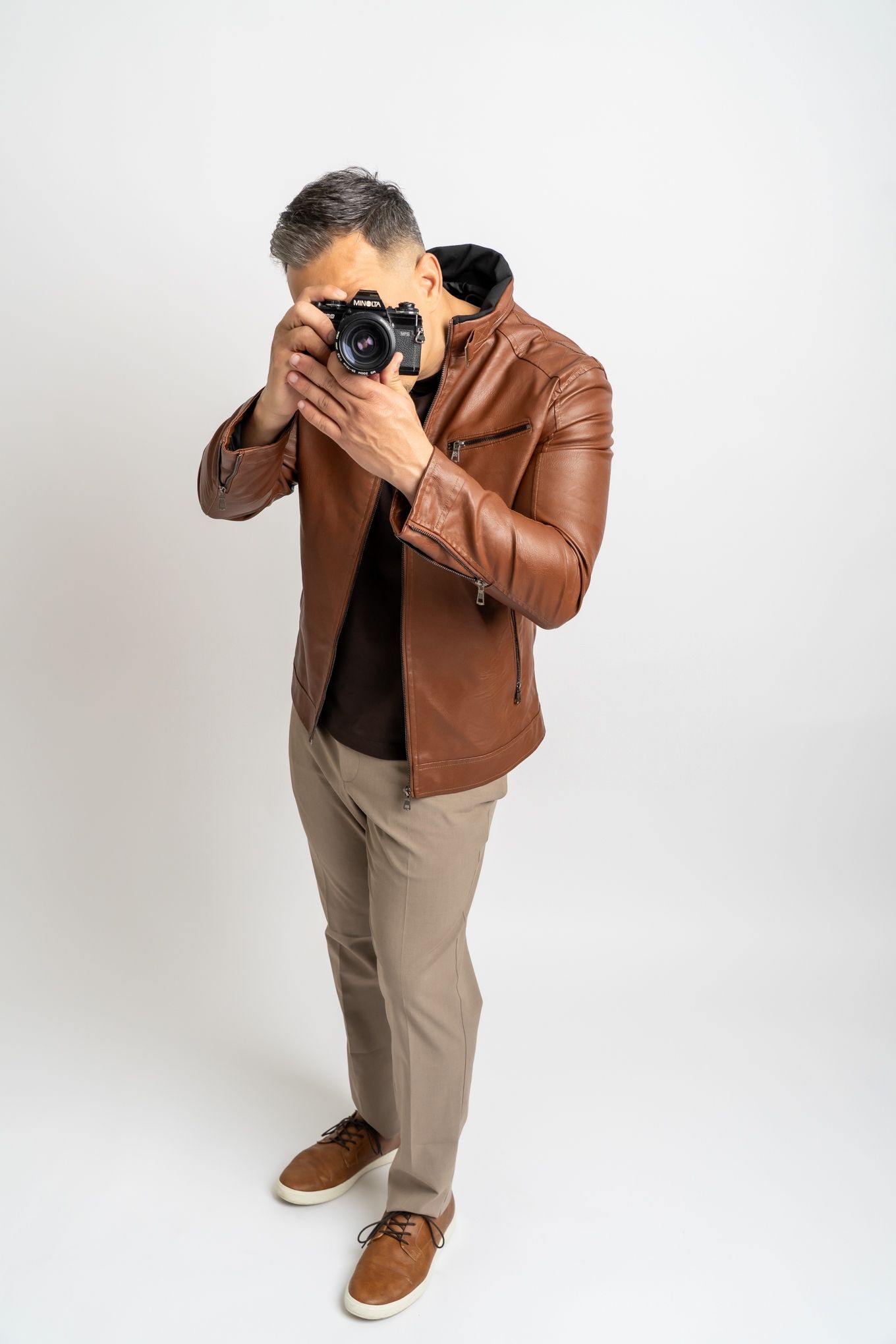 Professional photographer wearing brown leather jacket and khaki pants takes photo with vintage camera against white background.