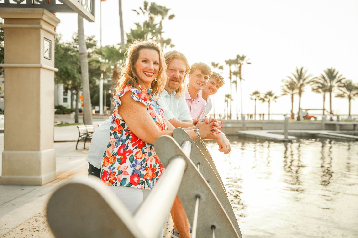 Group of people in casual attire smiling while leaning on a railing near a waterfront with palm trees in the background.
